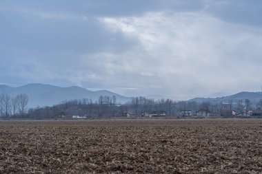 Ön planda kuru toprakla birlikte düz bir çiftlik alanı uzanır. Arka planda, alçak tepeler ve birkaç ev gri bulutların altında görülebilir. Sahne tipik bir kırsal alanı gösteriyor..