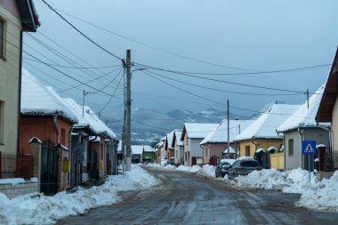Evler dar bir caddeyi çizerken kar zemini ve çatıları kaplıyor. Elektrik hatları üstten akıyor. Bir araba yolun kenarında kırsal bir yerde park halinde..