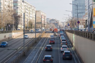 Şehirdeki geniş bir yolda arabalar sıraya dizildi. Arkaplanda binalar yükselirken otobüsler ve kamyonlar da mevcuttur. İnsanlar kaldırımlarda..
