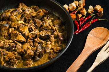 Tavuk ciğeri baharatlı bir tavada pişiriliyor ve küçük ekmek parçaları ve kırmızı biberle servis ediliyor. Mutfakta koyu renkli yemekler yapılır..