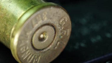 Görüntü, karanlık bir yüzeyde duran boş bir mermi kovanının yakın çekimini gösteriyor. Mermi kovanının ayrıntıları açık, işaretler görünüyor ve kullanılmaktan yıpranmış. Siyah arka planda, ateşli silahtan çıkan mermi kovanına yakından bakın.