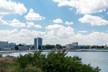 İnsanlar sahili ziyaret eder ve güneşli havanın tadını çıkarırlar. Bulutlar gökyüzünde süzülürken binalar kıyıya dizilir. Sahne, hareketli bir rıhtım alanı ve hareketlilik gösteriyor..