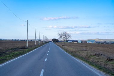 Kırsal kesimden geçen uzun bir yol var. Her iki tarafta da açık alanlar ve birkaç ağaç var. Açık mavi gökyüzünün altında küçük binalar belirir.