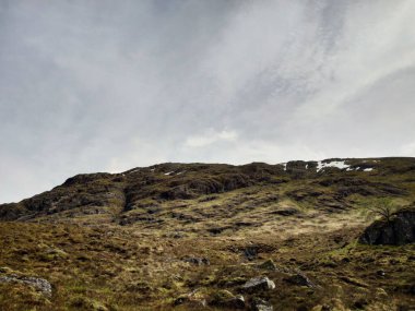 Bulutlu gökyüzünün altındaki kayalık yamaçta seyrek bitki örtüsü ve engebeli arazi