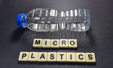 Mikro plastik kirliliğini gösteren kavramsal görüntü. Plastik atıkları sembolize eden, karanlık bir yüzeyde yatay olarak duran şeffaf bir plastik şişe. Altında, krem renkli karolar 'MICROPLASTICS' olarak heceleniyor, çevresel kaygıları vurguluyor.