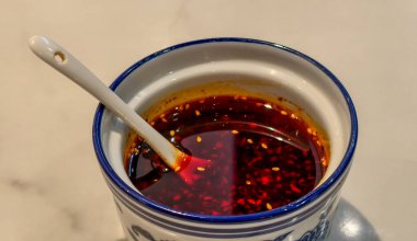 Mavi desenli geleneksel bir seramik kavanozda servis edilen görünen tanecikler ve tohumlarla acılı biber yağı. Beyaz bir seramik kaşık canlı kırmızı yağın içinde duruyor ve zengin dokusunu ve ısısını vurguluyor. Asya mutfağı için mükemmel..