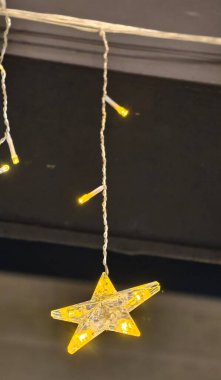 Parlak yıldız şekilli LED ışık, daha küçük silindirik ampullerle dekoratif bir sicimin parçası olan karanlık bir arkaplanda parlıyor. Şeffaf yıldız, sıcak sarı ışığı dağıtarak çarpıcı bir zıtlık ve kutlama dekorasyonu için neşeli bir atmosfer yaratır..