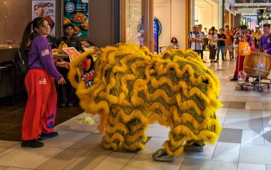 Sarı kostümlü sanatçıların olduğu bir alışveriş merkezinde aslan dansı gösterisi. Müşteriler ve çalışanlar izlerken bir davulcu büyük bir davul çalar..