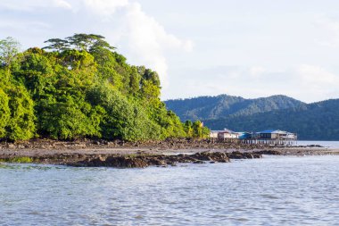 Geniş açılı bir manzara, sudaki sazlıklar üzerindeki geleneksel ahşap evleri, yemyeşil bir tepenin ve kayalık alçak gelgit plajının yanında gösteriyor.