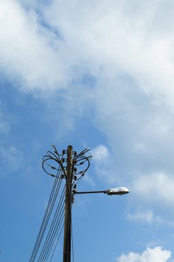 Dairesel tel düzenleyicili elektrik direği, yalıtımlı ve sokak lambası sabitleyicisi parçalı bulutlu gökyüzü altında, kentsel elektrik dağıtımını ve kamu aydınlatmasını temsil ediyor.