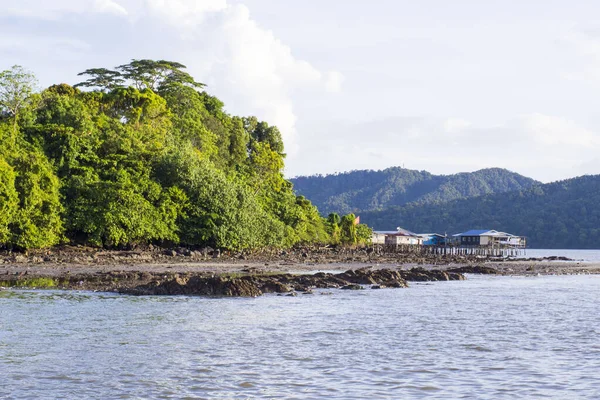 Geniş açılı bir manzara, sudaki sazlıklar üzerindeki geleneksel ahşap evleri, yemyeşil bir tepenin ve kayalık alçak gelgit plajının yanında gösteriyor.