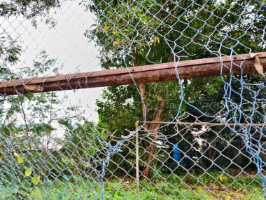 Paslı ve yırtık mavi kaplamalı tel örgüye yakın plan. Kırık kısımlar, arka planda yeşil ağaçlar ve bir yerleşim alanı olan ihmal veya zorla girildiğini gösteriyor.