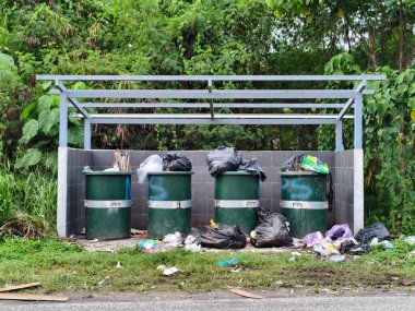 Modern gri metal bir sığınağın altına dizilmiş dört büyük yeşil plastik çöp kutusu. Siyah çöp torbaları çöp kutularının içine ve etrafına istiflenmiştir. Bu da tropikal bir bölgedeki kamu atık yönetim sistemini gösterir.