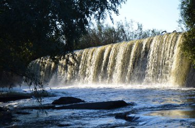 Ağaçlar ve bereketli doğayla çevrili alçak bir barajın üzerinde çağlayan manzaralı şelale ön planda akan nehir suyuyla sıcak güneş ışığıyla aydınlatılıyor..