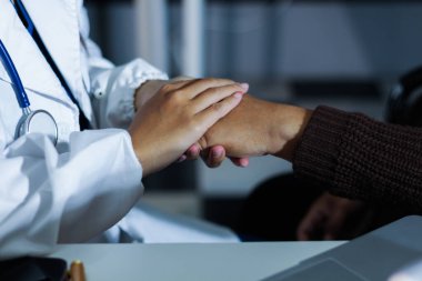 Hastane yatağında yatan kıdemli hasta genç kadın hemşireden teselli edici bir dokunuş alıyor. Elleri nazikçe birbirine kenetlenmiş, tıbbi bakım ayarları görülebilir.