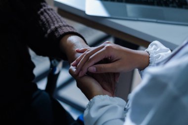 Hastane yatağında yatan kıdemli hasta genç kadın hemşireden teselli edici bir dokunuş alıyor. Elleri nazikçe birbirine kenetlenmiş, tıbbi bakım ayarları görülebilir.
