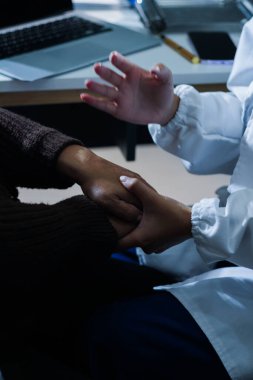 Hastane yatağında yatan kıdemli hasta genç kadın hemşireden teselli edici bir dokunuş alıyor. Elleri nazikçe birbirine kenetlenmiş, tıbbi bakım ayarları görülebilir.