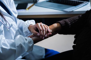 Hastane yatağında yatan kıdemli hasta genç kadın hemşireden teselli edici bir dokunuş alıyor. Elleri nazikçe birbirine kenetlenmiş, tıbbi bakım ayarları görülebilir.