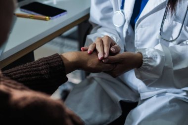 Hastane yatağında yatan kıdemli hasta genç kadın hemşireden teselli edici bir dokunuş alıyor. Elleri nazikçe birbirine kenetlenmiş, tıbbi bakım ayarları görülebilir.