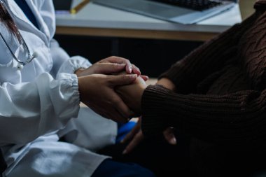 Hastane yatağında yatan kıdemli hasta genç kadın hemşireden teselli edici bir dokunuş alıyor. Elleri nazikçe birbirine kenetlenmiş, tıbbi bakım ayarları görülebilir.