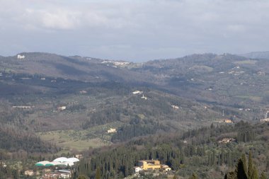 Orman Havzası boyunca zeytin kemerleri ve selvi hatlarıyla dağılmış uzak villalar, sessiz gün ortası ışığı ve katmanlı tepeler kırsal araziyi öneriyor
