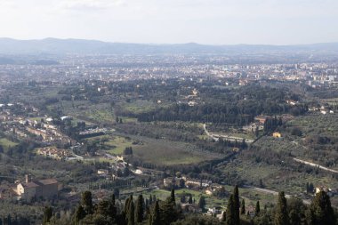 Ön planda zeytin ağaçları ve üzüm bağları olan Vadi Tarlaları ve Villalar temiz ışığın altında yuvarlanan tepelerin üzerinde görünen uzak Floransa silueti..