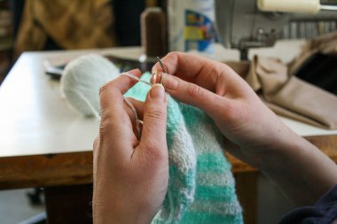 Örgü yaparken bir kadının ellerini gösteren yakın plan bir fotoğraf, örgü iğneleriyle dikkatlice çalışan bir iplik. Görüntü, el işçiliğinin detaylarını yakalıyor, doku, hareket ve örgü örmenin sakin, yaratıcı ritmine odaklanıyor..