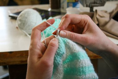 Örgü yaparken bir kadının ellerini gösteren yakın plan bir fotoğraf, örgü iğneleriyle dikkatlice çalışan bir iplik. Görüntü, el işçiliğinin detaylarını yakalıyor, doku, hareket ve örgü örmenin sakin, yaratıcı ritmine odaklanıyor..