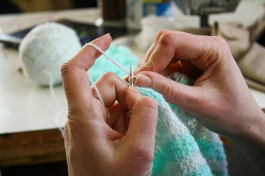 Örgü yaparken bir kadının ellerini gösteren yakın plan bir fotoğraf, örgü iğneleriyle dikkatlice çalışan bir iplik. Görüntü, el işçiliğinin detaylarını yakalıyor, doku, hareket ve örgü örmenin sakin, yaratıcı ritmine odaklanıyor..
