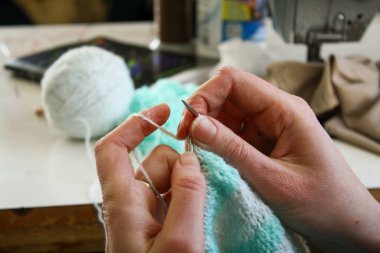 Örgü yaparken bir kadının ellerini gösteren yakın plan bir fotoğraf, örgü iğneleriyle dikkatlice çalışan bir iplik. Görüntü, el işçiliğinin detaylarını yakalıyor, doku, hareket ve örgü örmenin sakin, yaratıcı ritmine odaklanıyor..