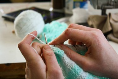 Örgü yaparken bir kadının ellerini gösteren yakın plan bir fotoğraf, örgü iğneleriyle dikkatlice çalışan bir iplik. Görüntü, el işçiliğinin detaylarını yakalıyor, doku, hareket ve örgü örmenin sakin, yaratıcı ritmine odaklanıyor..