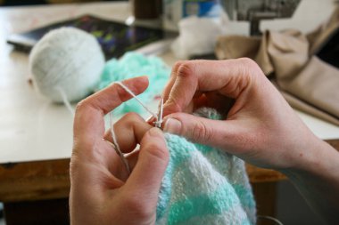Örgü yaparken bir kadının ellerini gösteren yakın plan bir fotoğraf, örgü iğneleriyle dikkatlice çalışan bir iplik. Görüntü, el işçiliğinin detaylarını yakalıyor, doku, hareket ve örgü örmenin sakin, yaratıcı ritmine odaklanıyor..