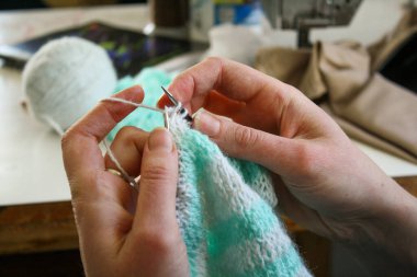 Örgü yaparken bir kadının ellerini gösteren yakın plan bir fotoğraf, örgü iğneleriyle dikkatlice çalışan bir iplik. Görüntü, el işçiliğinin detaylarını yakalıyor, doku, hareket ve örgü örmenin sakin, yaratıcı ritmine odaklanıyor..
