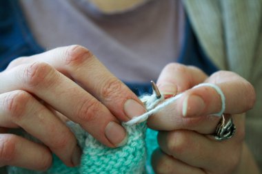 Örgü yaparken bir kadının ellerini gösteren yakın plan bir fotoğraf, örgü iğneleriyle dikkatlice çalışan bir iplik. Görüntü, el işçiliğinin detaylarını yakalıyor, doku, hareket ve örgü örmenin sakin, yaratıcı ritmine odaklanıyor..
