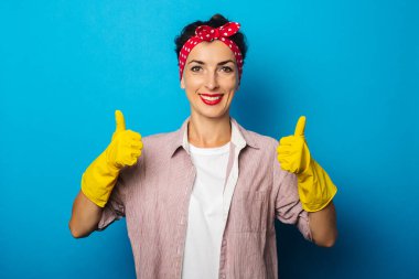 Gülümseyen genç kadın mavi arka planda temizlik eldivenlerinde baş parmak işareti gösteriyor.