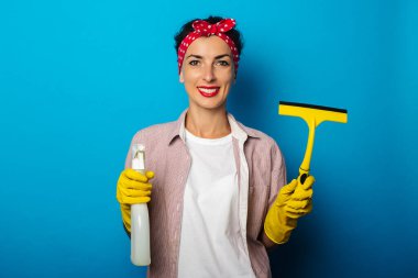Eldivenli gülümseyen genç kadın mavi arka plandaki pencereleri temizlemek için sprey ve kazıyıcı tutuyor.