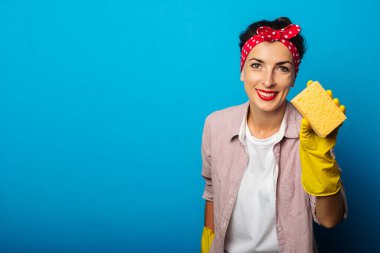 Mavi arka planda bir sünger tutan eldivenli, saç bandı takmış gülümseyen genç bir kadın.