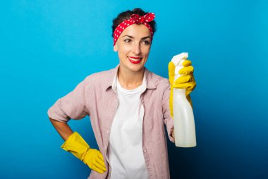 Elinde sprey tutan temizlik eldivenleriyle gülümseyen genç kadın mavi arka planda ona bakıyor.