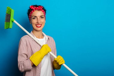 Gülümseyen, temizlik eldivenli, mavi arka planda temizlik fırçası tutan genç bir kadın.