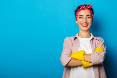 Saçı bandajlı, mavi arka planda eldivenlerini temizlerken ellerini çaprazlayan arkadaş canlısı genç bir kadın.