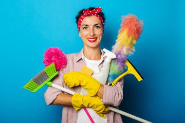Sarı eldivenli gülümseyen genç kadın mavi arka planda temizlik aksesuarları tutuyor.