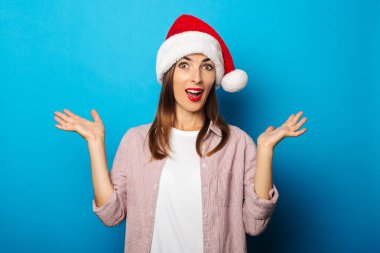 Mavi arka planda Noel Baba şapkası takan tişörtlü, neşeli genç bir kadın.