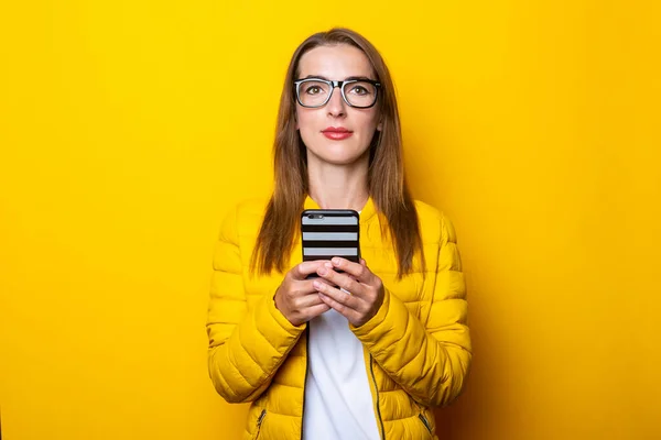 Young woman in a yellow jacket thoughtfully holding a phone on a yellow background