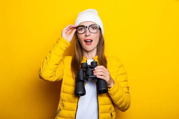 Surprised young woman in yellow jacket with binoculars on yellow background