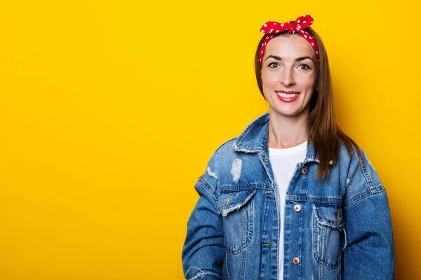 Smiling young woman in hair band, in denim jacket on yellow background