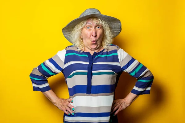 Surprised old woman in dress and hat holds her hands at the waist on a yellow background.