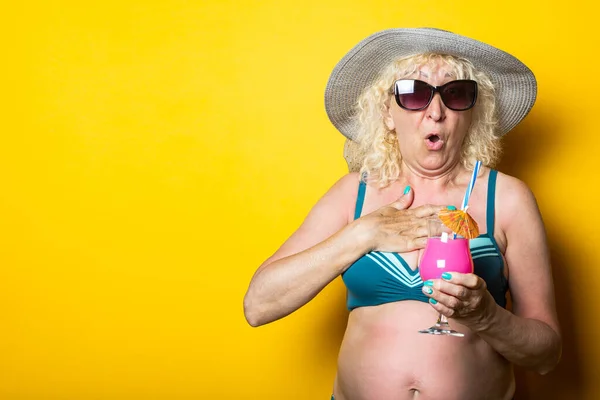 Old woman in a swimsuit with a hat holding a cocktail in surprise on a yellow backgroun