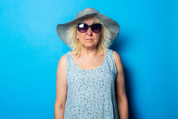 Old woman in dress, hat and sunglasses on blue background