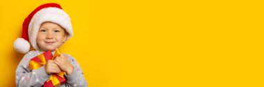 Noel şapkalı çocuk sarı arka planda bir hediye tutuyor. Noel hediyeleri, Noel sabahı. Pankart.
