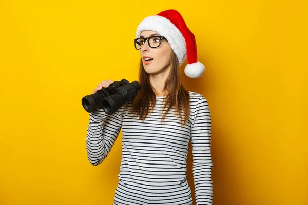 Mujer Joven Sombrero Navidad Sonriendo Sosteniendo Binoculares Sobre Fondo Amarillo — Foto de Stock